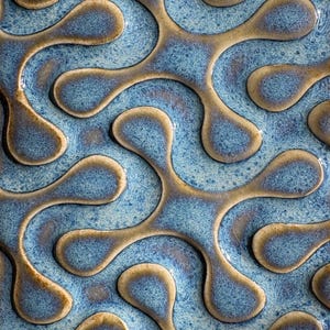 May include: A close-up of a ceramic tile with a repeating pattern. The design features interconnected, organic shapes in shades of blue and brown. The surface has a glossy finish, with a speckled texture in the blue areas.