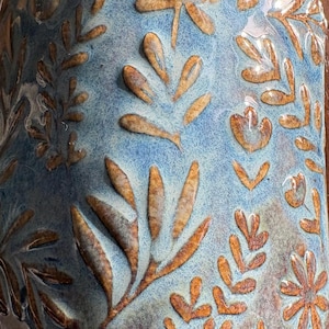 May include: Close-up of a ceramic vase with a blue and brown floral design. The vase has a textured surface with raised leaf and flower patterns in shades of brown. The background is a mottled blue, creating a rustic aesthetic.