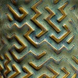 May include: A ceramic surface featuring a raised, three-dimensional maze pattern in a glossy golden-brown finish. The recessed areas between the geometric lines are filled with a mottled, dark teal and sage green glaze with a smooth texture.
