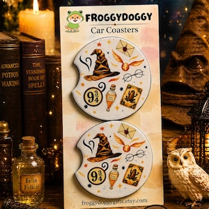 May include: Two white car coasters with a magical theme. Each coaster features a brown sorting hat, gold snitch, glasses, and a potion bottle. The coasters also have the text "9 3/4" and "Felix Felicis". The brand name "FROGGYDOGGY" is at the top.