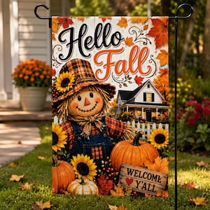 May include: A decorative garden flag featuring a scarecrow wearing a plaid hat and shirt with overalls, surrounded by sunflowers and pumpkins. The text reads Hello Fall and Welcome Y'all. A white farmhouse is visible in the background with autumn leaves.