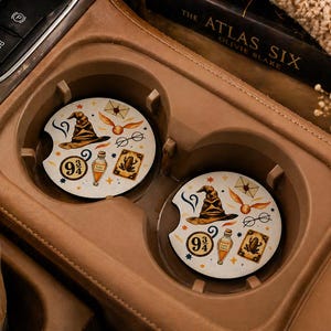 May include: Two round car coasters with a cream background and a wizarding theme. Each coaster features a sorting hat, golden snitch, glasses, potion bottle, and a 9 3/4 symbol. The coasters are in a car's cup holder.