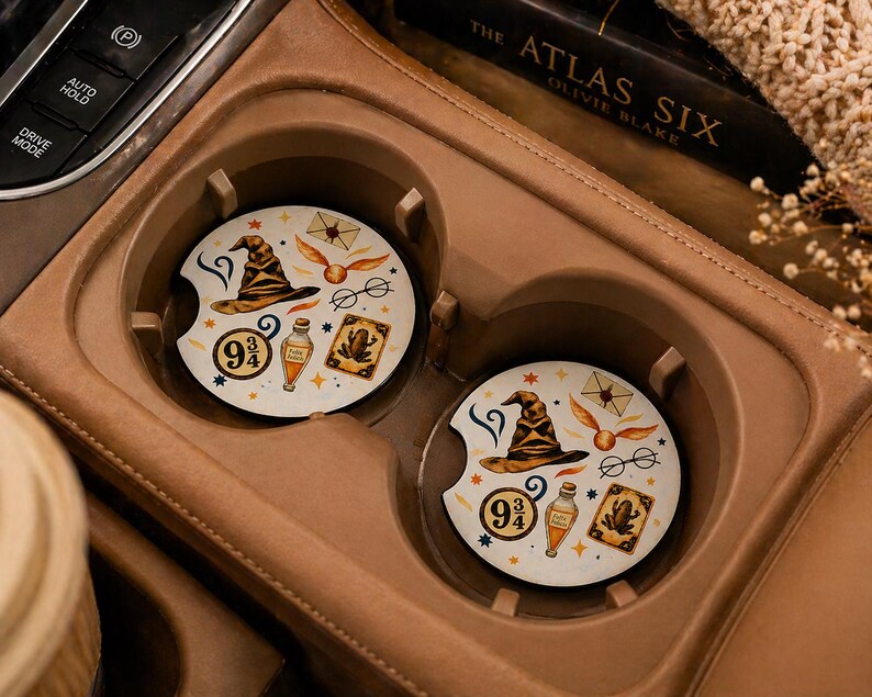 May include: Two round car coasters with a cream background and a wizarding theme. Each coaster features a sorting hat, golden snitch, glasses, potion bottle, and a 9 3/4 symbol. The coasters are in a car's cup holder.