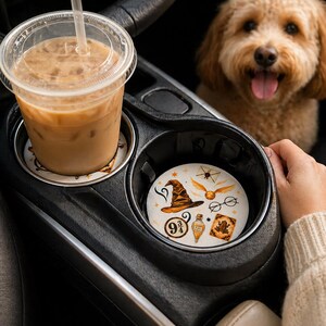 May include: A car cup holder with two coasters. One coaster has a coffee drink on top. The other coaster has a wizard hat, glasses, potion bottle, and other magical themed images. A dog is in the background.