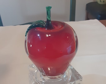 May include: A red glass apple sculpture with a green stem and leaf, sitting on a clear glass square base.