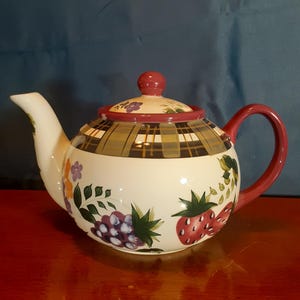 May include: A white ceramic teapot with a red handle and lid. The teapot is decorated with a plaid pattern on the lid and a design of grapes and strawberries. The spout is white and the handle is red.