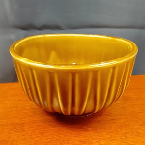 May include: A ceramic bowl in a warm, golden-brown hue, featuring a ribbed design on the exterior. The bowl has a rounded form and is placed on a wooden surface. The interior is smooth and glossy.