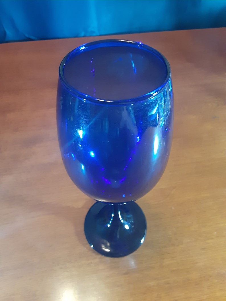 May include: A cobalt blue glass goblet with a wide bowl and a stem. The goblet is empty and sitting on a wooden surface.