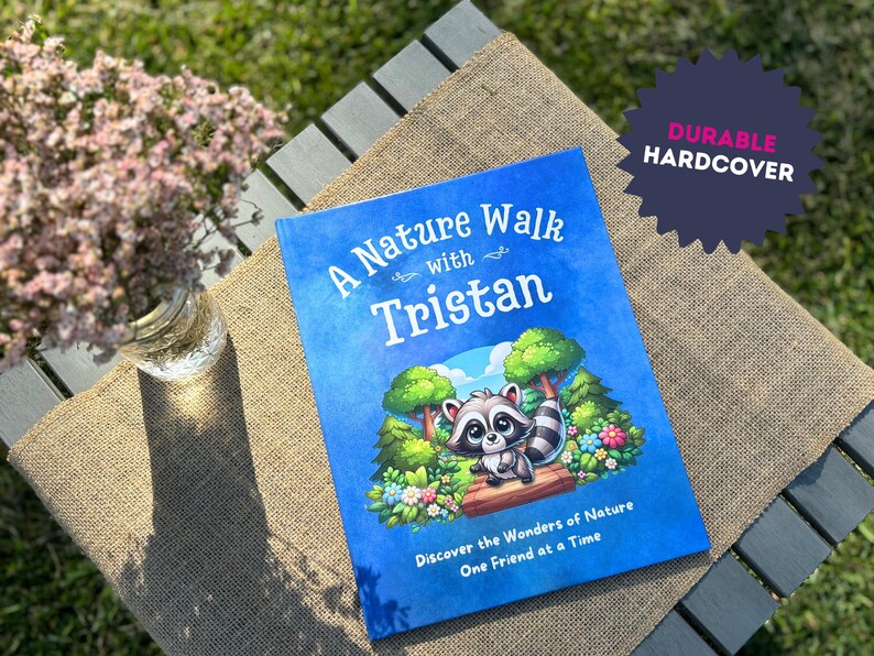 May include: A blue hardcover book with the title "A Nature Walk with Tristan" and an illustration of a raccoon in a forest setting. The book is on a brown burlap surface.