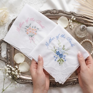 Könnte beinhalten: Zwei weiße, bestickte Taschentücher. Eines zeigt einen rosa-orangenen Blumenstrauß mit dem Text "Mother of the Bride", das andere blaue Blumen. Beide haben Spitzensäume und liegen auf einem silbernen Tablett.