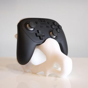 May include: A black Nintendo Switch Pro controller resting on a white, skull-shaped 3D-printed stand. The controller features standard buttons, two analogue sticks, and the Nintendo Switch logo printed in white on the top centre of the device.
