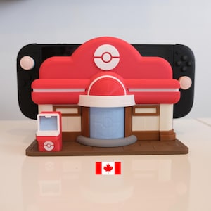 May include: A red, white, and brown 3D-printed model of a building with a matching vending machine. The model is in front of a black Nintendo Switch Lite. The building has a white and red design with a circular logo on top.