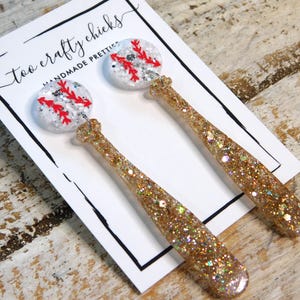 Peut inclure: Une paire de boucles d'oreilles en forme de batte de baseball avec des paillettes dorées et des breloques de baseball blanches avec des coutures rouges. Les boucles d'oreilles sont présentées sur une carte blanche avec le texte "too crafty chicks handmade pretties".