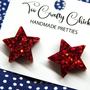May include: A pair of red glitter star-shaped earrings on a white card with the text "Too Crafty Chicks HANDMADE PRETTIES". The earrings are set against a blue and white polka dot background.