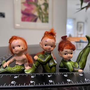 Könnte beinhalten: Drei Vintage-Keramikfiguren: eine Meerjungfrau mit grünem Schwanz und zwei Figuren in grünen Outfits. Die Figuren haben rotes Haar und sind in verschiedenen Positionen dargestellt. Die Meerjungfrau sitzt, eine Figur sitzt und die andere macht einen Handstand.