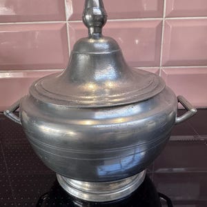 May include: A large, silver pewter serving bowl with a lid and two handles. The bowl has a rounded body and a raised base. The lid has a finial in the shape of an acorn.