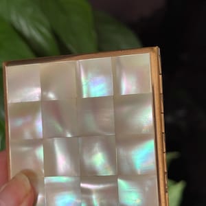 Può includere: Un compatto quadrato vintage con una cornice color oro e un design a mosaico di madreperla sul coperchio. Il mosaico è composto da piccole tessere quadrate che riflettono colori iridescenti. Il compatto è tenuto in mano, con uno sfondo verde sfocato.