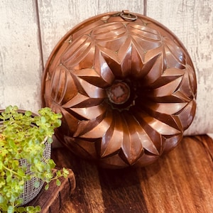 May include: A vintage copper bundt pan with a decorative starburst pattern. The pan has a ring for hanging and is displayed on a wooden surface. The copper has a warm, reddish-brown hue, suggesting age and use.