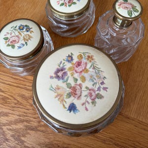 Puede incluir: Conjunto de cuatro frascos de vidrio vintage con tapas bordadas con flores. Los frascos son de vidrio transparente con un diseño facetado. Las tapas son blancas con bordados florales coloridos y ribetes dorados.