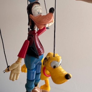 Könnte beinhalten: Ein Vintage-Marionetten-Set von Goofy und Pluto. Goofy trägt einen blauen Hut, eine rote Weste und eine blaue Hose. Pluto ist gelb mit einem roten Halsband. Beide Marionetten sind an schwarzen Fäden aufgehängt.