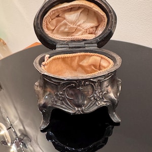 May include: An antique silver-toned jewelry box with an oval shape and ornate floral detailing. The box is open, revealing a peach-colored fabric lining. The box sits on a reflective surface.