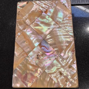 May include: Rectangular mother-of-pearl box with a woven pattern of iridescent squares. The box has a light brown frame and a clasp. The iridescent squares reflect colours of pink, green, and gold.