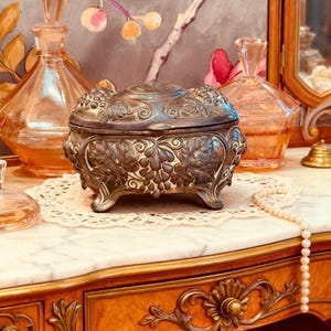 May include: An antique silver-toned jewelry box with ornate leaf and grape detailing. The box sits on a lace doily atop a marble-topped wooden vanity. Two peach-colored perfume bottles are visible in the background, along with a string of pearls.
