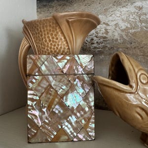 May include: A rectangular box covered in iridescent diamond-shaped mother-of-pearl tiles sits between two tan ceramic fish-shaped pitchers. The box features a hinged lid and a shimmering, multi-colored surface with pink, green, and gold tones.