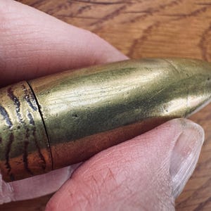 May include: A brass bullet-shaped object with a textured, dark band at the base. The bullet is held between fingers, with a wooden surface in the background. The object has a shiny, metallic appearance.