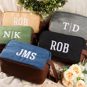 May include: A collection of personalized toiletry bags in various colors, including tan, green, blue, black, and gray. Each bag features embroidered initials in white. The bags have brown leather-like bottoms and handles.