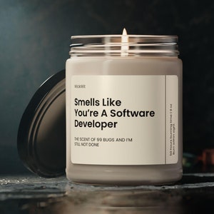 Könnte beinhalten: Eine beige Kerze in einem Glasgefäß mit schwarzem Deckel. Das Etikett lautet "Smells Like You're A Software Developer" und "The scent of 99 bugs and I'm still not done".