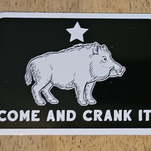 以下が含まれることがあります： 白と黒の星とイノシシのイラストが描かれたステッカー。ステッカーには白い文字で「COME AND CRANK IT」と書いてあります。