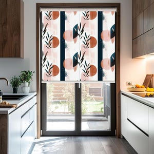 May include: A white window blind with a geometric pattern featuring pink, orange, and blue abstract shapes and leaves. The blind is partially rolled up, revealing a view of a green lawn outside the window.