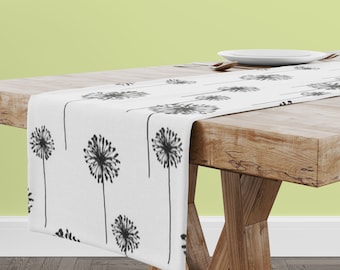 May include: White table runner with a black dandelion print. The runner is on a wooden table with a white plate and silverware.