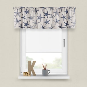 May include: A valance curtain with a cream background and a pattern of blue starfish. The curtain is mounted above a white window. A shelf below the window holds decorative items, including a gold ring, a wooden letter "K", and a gray mug with pencils.