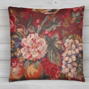 May include: A decorative pillow with a floral pattern. The pillow features a burgundy background with a repeating pattern of large flowers in shades of red, pink, and white, along with green leaves and other floral elements. The pillow is square.