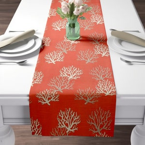 May include: A coral-colored table runner with a white coral design. The runner is made of a textured fabric and has a subtle sheen.