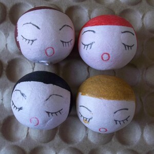 May include: Four round, white, paper mache ornaments with painted faces. Each ornament has closed eyes, a red mouth, and a different colored hair: brown, red, black, and gold.