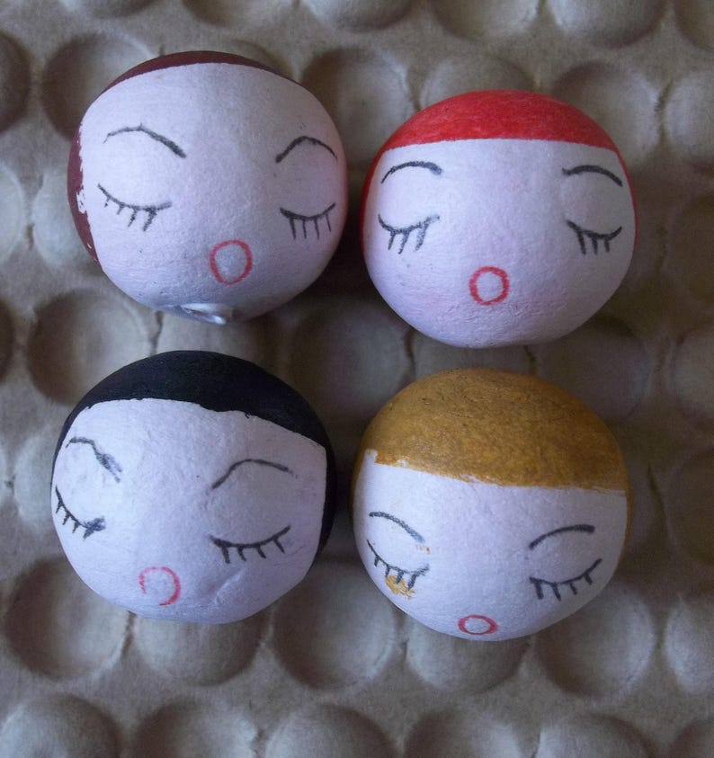 May include: Four round, white, paper mache ornaments with painted faces. Each ornament has closed eyes, a red mouth, and a different colored hair: brown, red, black, and gold.