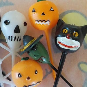 May include: A collection of vintage Halloween-themed decorations. Includes a white skull, two orange jack-o'-lanterns, a green witch, and a black cat. Each figure is mounted on a thin, long stem, perfect for cake toppers or party favors.