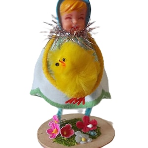 May include: A vintage Easter figurine of a smiling doll wearing a blue bonnet and a white dress with a yellow chick. The doll stands on a round base with pink, red, and white flowers and green moss.