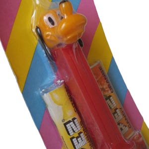 May include: A vintage PEZ dispenser featuring a yellow cartoon character head with black ears and eyes, and a red dispenser body. The candy is packaged in a colorful striped cardboard backing with the word "BONBONS" visible.