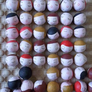 May include: A collection of small, round, painted wooden ornaments with closed eyes and various facial expressions. The ornaments are arranged in rows on a brown textured surface.