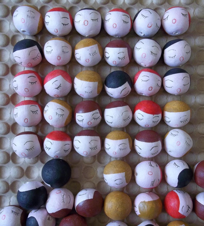 May include: A collection of small, round, painted wooden ornaments with closed eyes and various facial expressions. The ornaments are arranged in rows on a brown textured surface.
