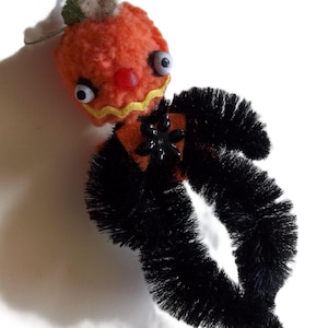 May include: A handcrafted figure made from orange pom-poms and black pipe cleaners. The head features two googly eyes, a small red nose, and a yellow zigzag mouth. A black plastic flower embellishment is attached to the center of the body.