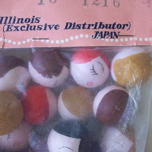 May include: A close-up of a bag of small, round, painted wooden dolls with various facial expressions. The dolls are in a clear plastic bag with a pink and white label that reads "Illinois (Exclusive Distributor) JAPAN".