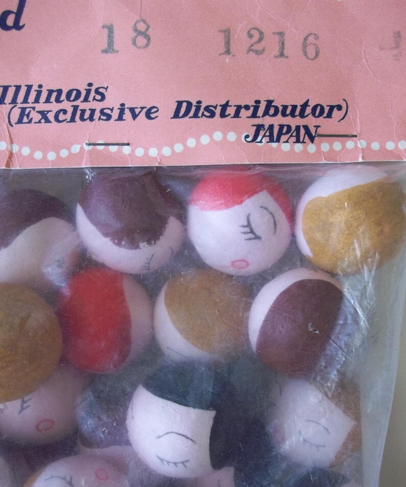 May include: A close-up of a bag of small, round, painted wooden dolls with various facial expressions. The dolls are in a clear plastic bag with a pink and white label that reads "Illinois (Exclusive Distributor) JAPAN".