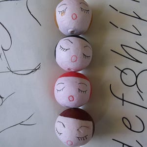 May include: Four round, white, paper mache ornaments with painted faces. Each ornament has closed eyes, a red mouth, and a different colored top. The top colors are brown, black, red, and yellow.