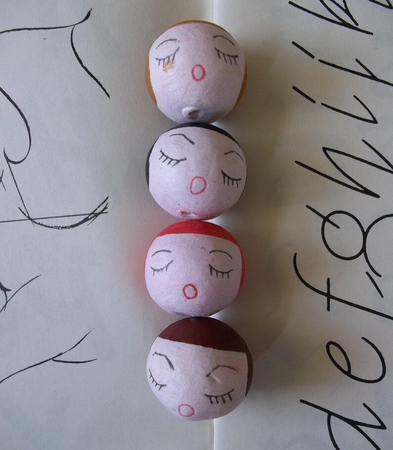 May include: Four round, white, paper mache ornaments with painted faces. Each ornament has closed eyes, a red mouth, and a different colored top. The top colors are brown, black, red, and yellow.
