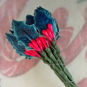 May include: A bouquet of teal blue ceramic holly leaves with pink berry accents. The leaves are arranged in a fan shape and the berries are clustered together in the center. The bouquet is wrapped in green paper and tied with wire.